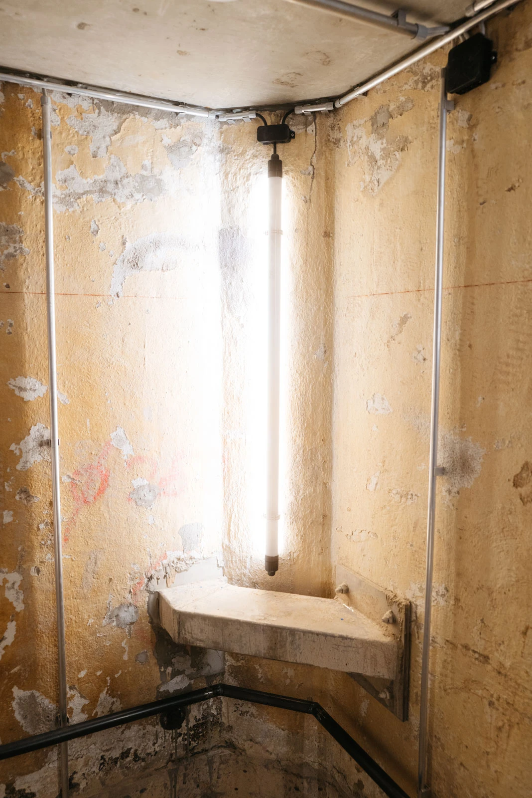 Vertical fluorescent light mounted on a peeling beige plaster wall in a basement utility corner, with a concrete shelf and exposed pipes.