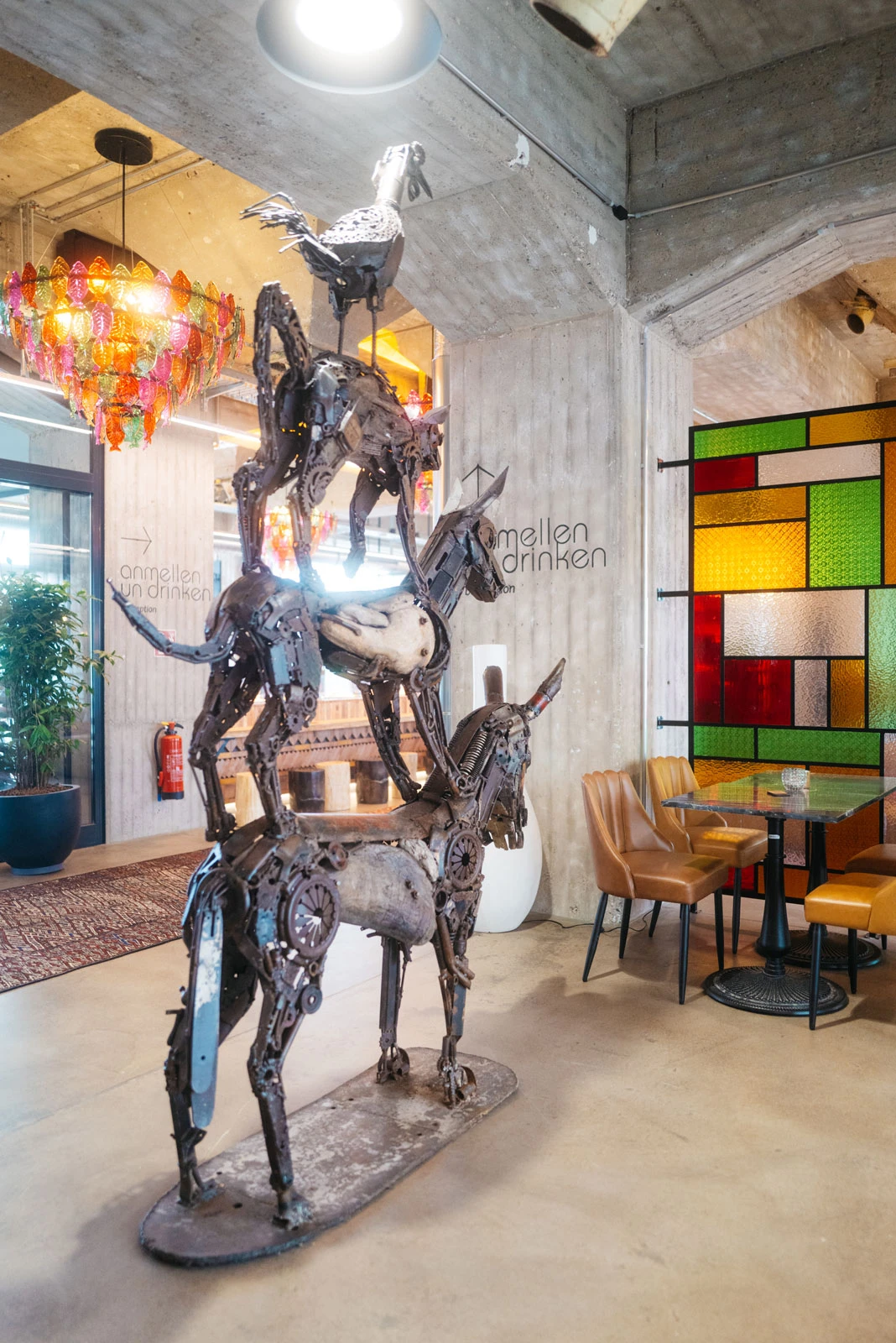 Scrap-metal sculpture of a horse with a bird perched on its back, in a modern industrial cafe with a colorful stained-glass wall.