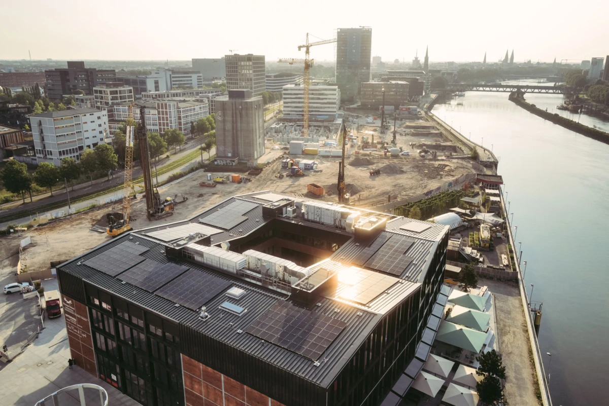 Aerial view of a riverfront construction site with cranes and machinery beside a modern glass-clad building with solar panels on its roof.