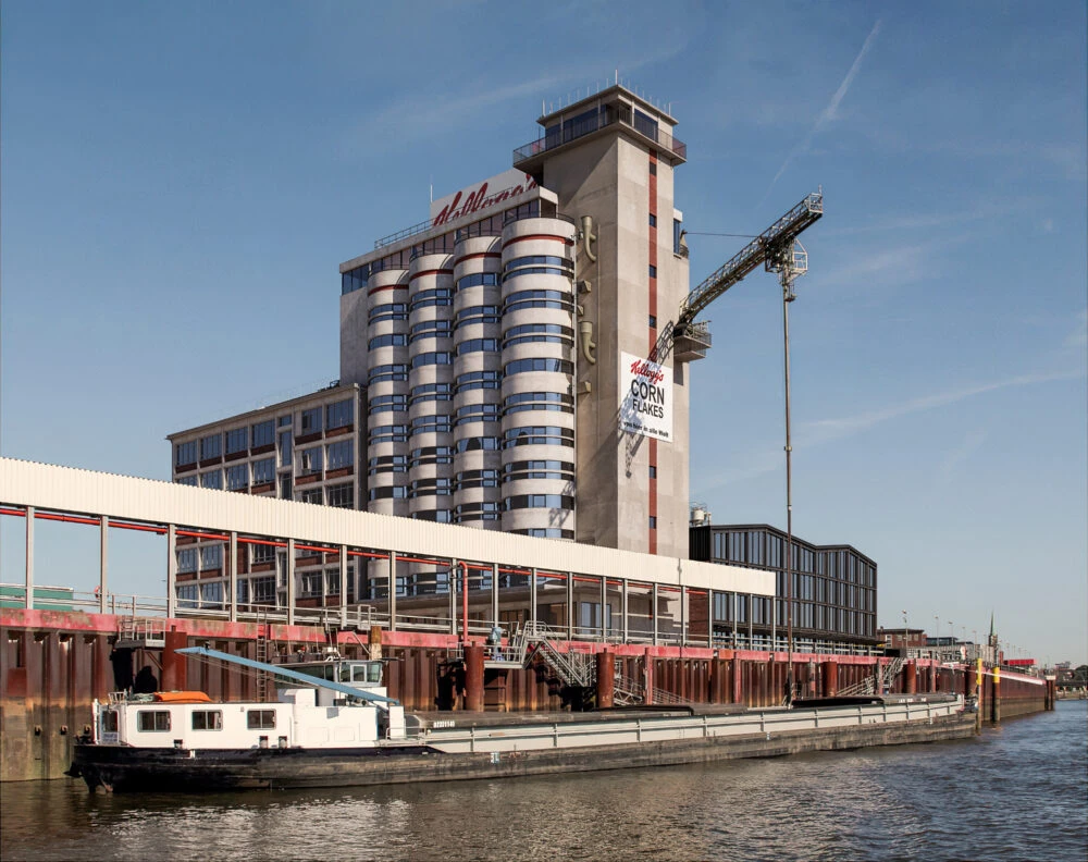 Kellogg's Corn Flakes factory on the riverfront with tall cylindrical silos, loading crane, and a docked barge.
