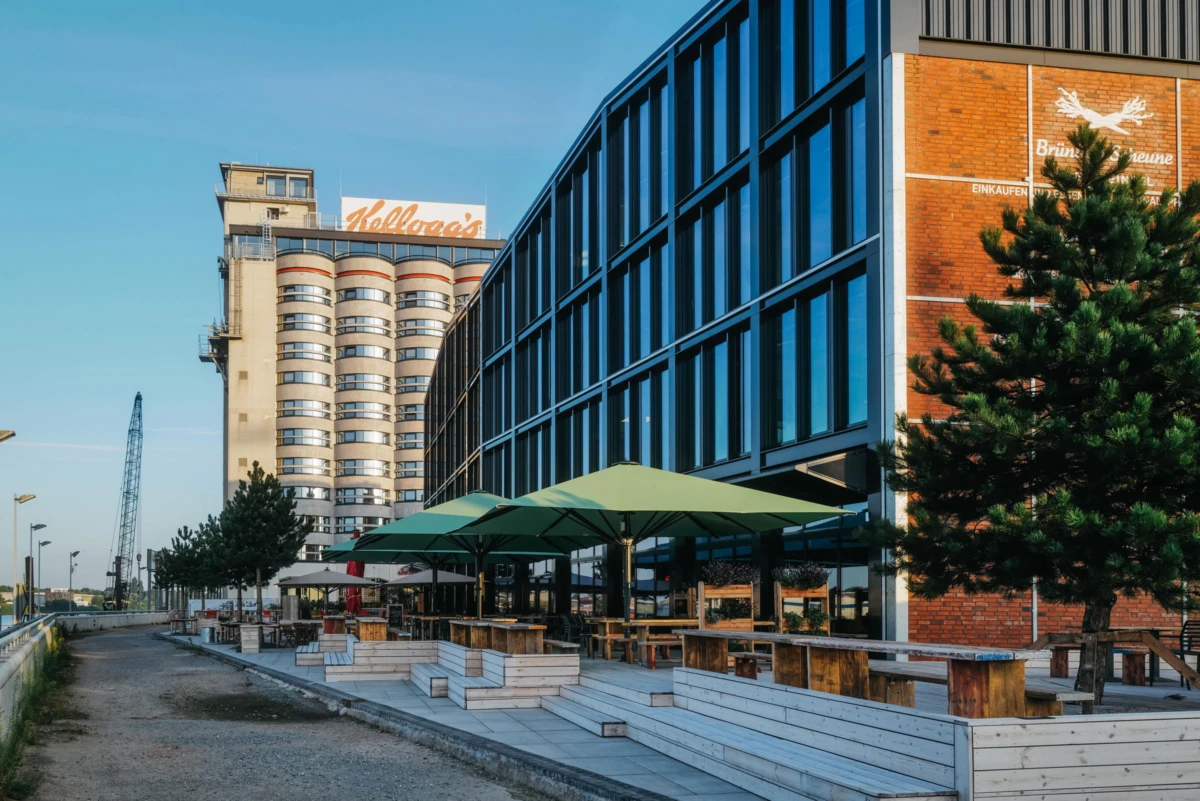 Empty outdoor cafe area with wooden benches and green umbrellas beside a glass-fronted office building along a riverside promenade.