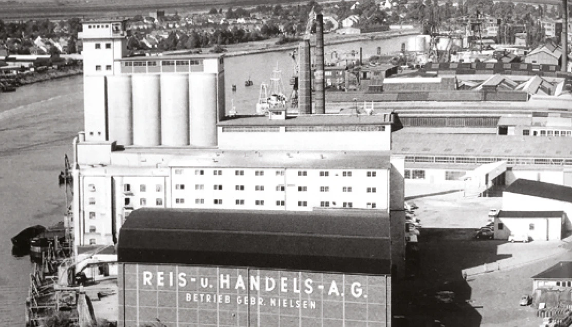 Portside industrial complex with tall silos and a curved-front warehouse displaying 'REIS-u.HANDELS-A.G.' along the waterfront.