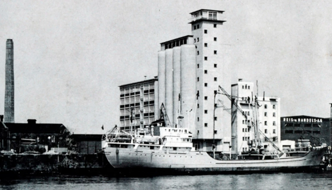 Cargo ship moored at an industrial harbor beside grain silos and tall buildings.