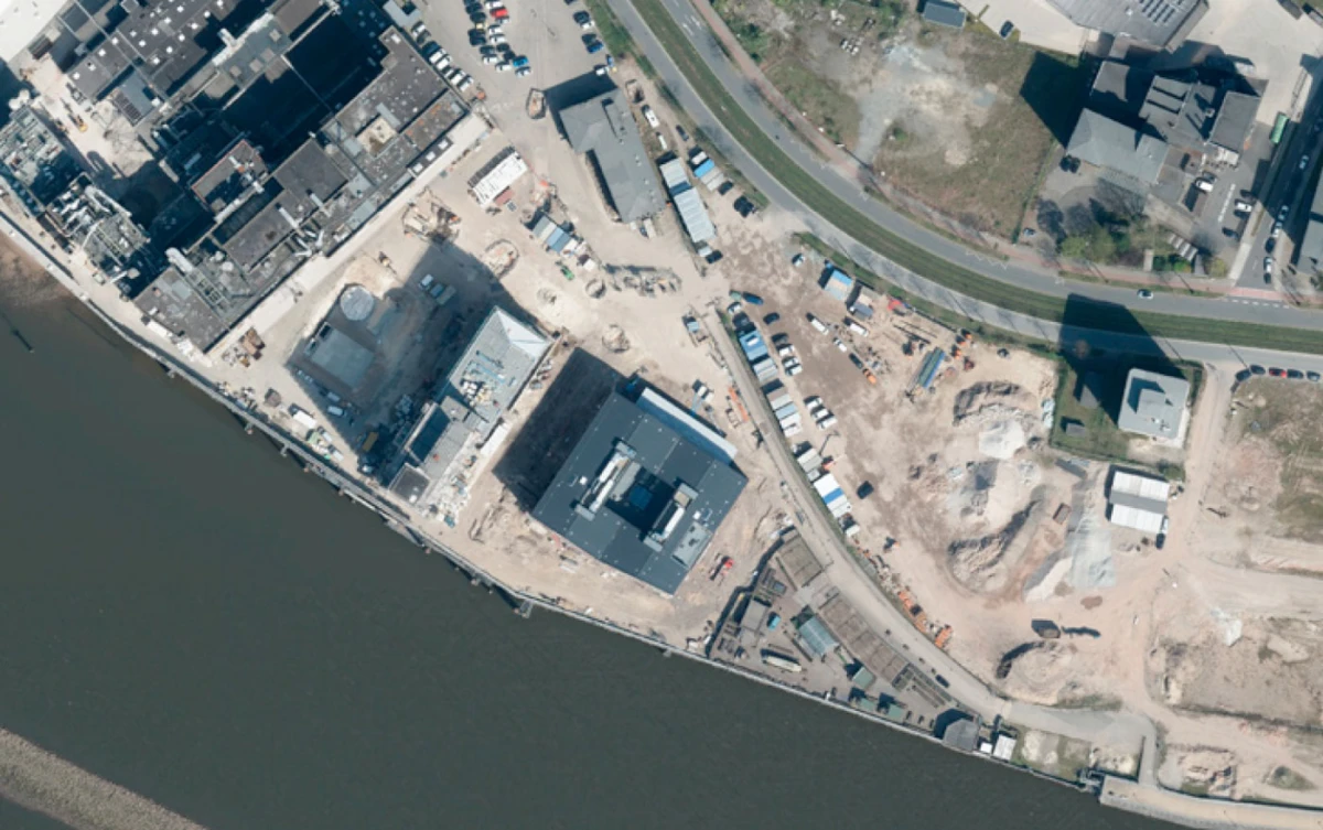 Aerial view of a riverfront construction site with partially built buildings, heavy equipment, and cleared ground along a curved road.