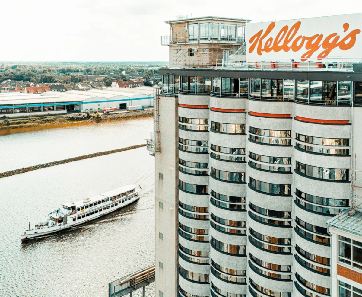 Cylindrical high-rise with rounded balconies along a river; Kellogg's sign atop; a white passenger boat sails nearby.