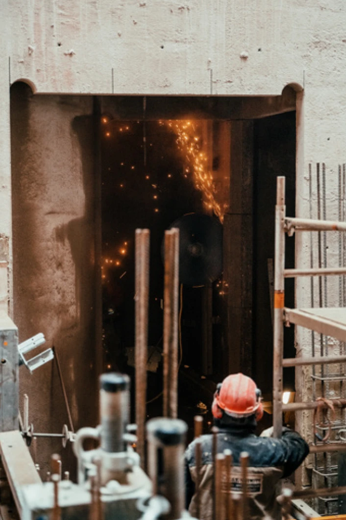 Construction worker in a red hard hat welding at a doorway; sparks fly inside a concrete building site with scaffolding nearby.