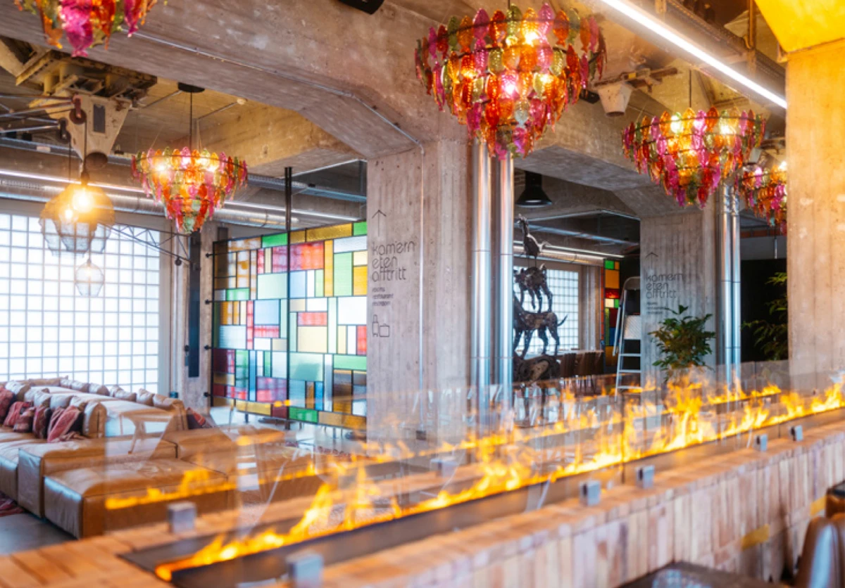 Industrial lounge with leather sectional sofas, multicolored stained-glass wall, hanging chandeliers, and a flame-lit bar counter.