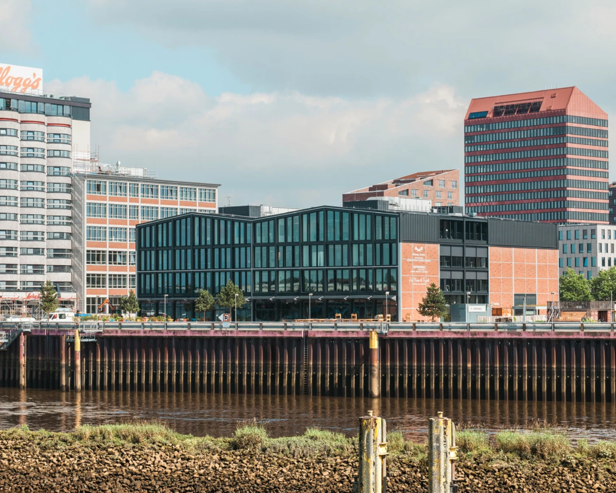 Glass-fronted office building along a riverfront with brick accents, framed by an urban skyline and a pier in the foreground.