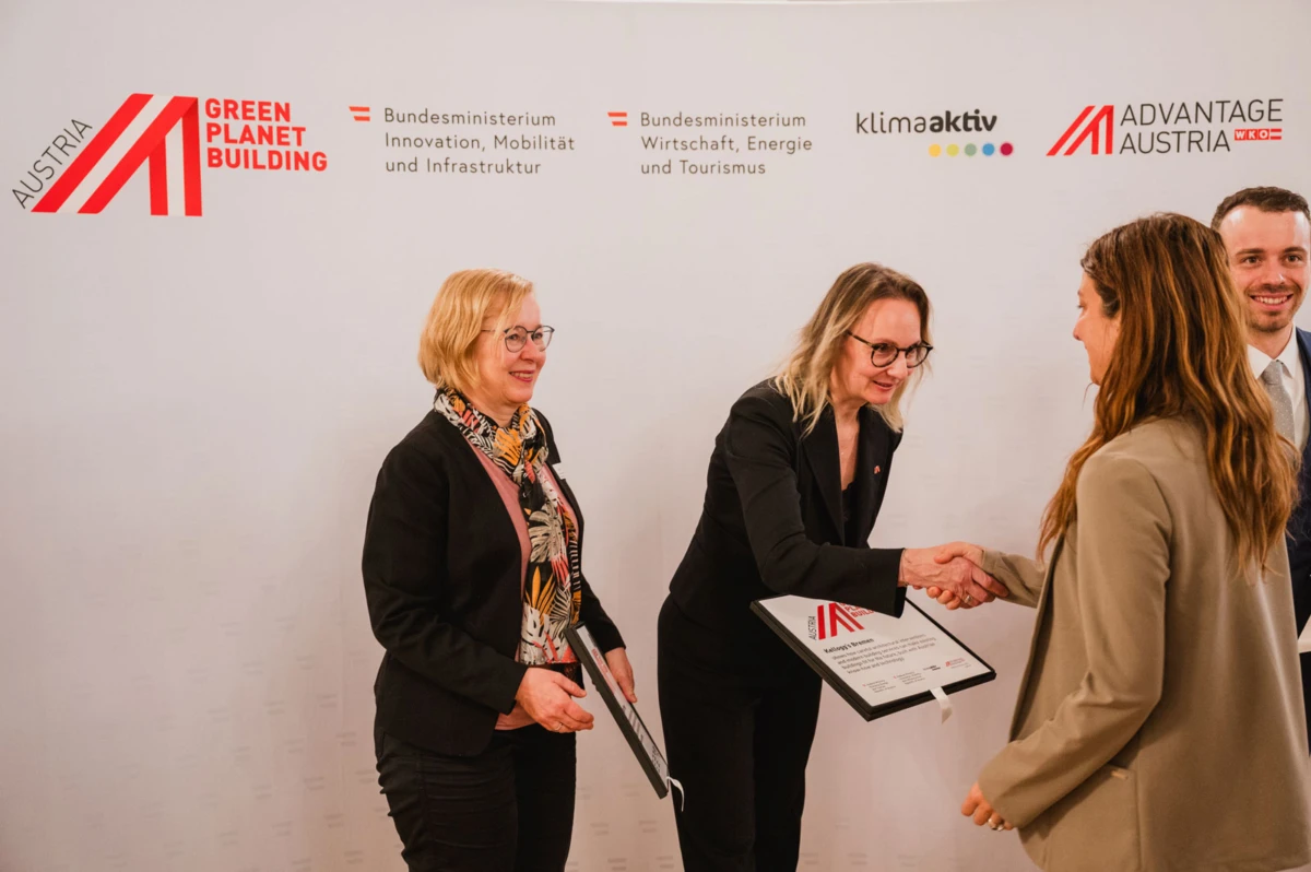 Two women shake hands during a formal ceremony; one holds a framed certificate as a third woman stands nearby; backdrop shows Austrian ministry logos.
