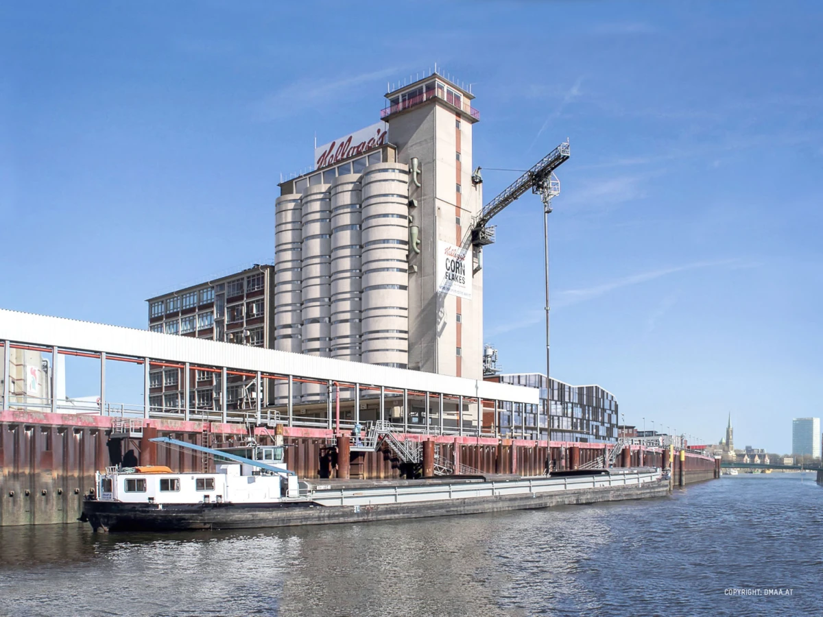 Industrial riverfront with stacked cylindrical silos, a tall tower, crane, and a barge along the pier under a blue sky.
