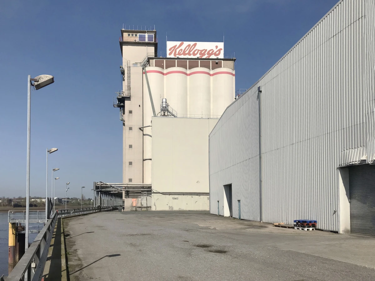Kellogg's cereal plant with tall silos and a white industrial building, empty loading area beside a waterfront walkway.