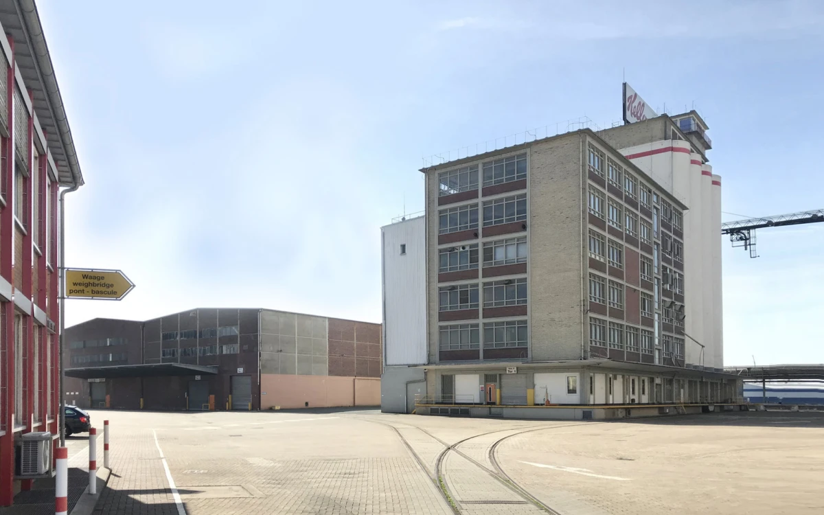 Industrial complex with a tall brick-and-concrete building, loading platform, and curved railway tracks across an open yard under a blue sky.