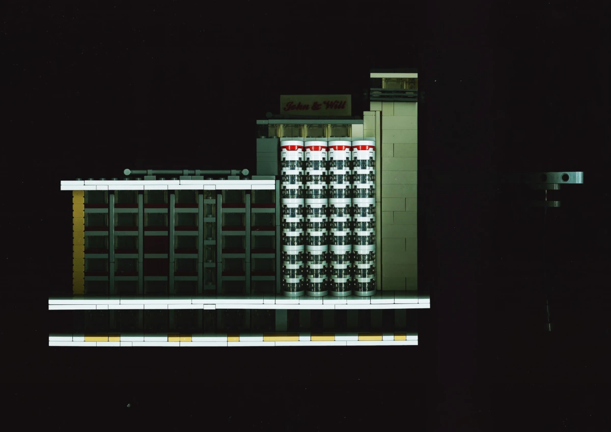 LEGO architectural model of a modern office building with central cylindrical stair towers and a 'John & Will' sign, set against a black background.