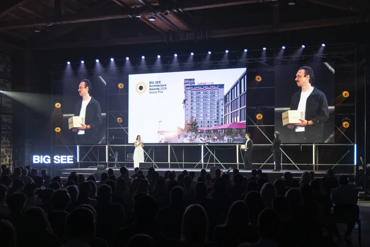 Presenter in white on stage at the Big See architectural awards ceremony, holding a box; large screens display a building.