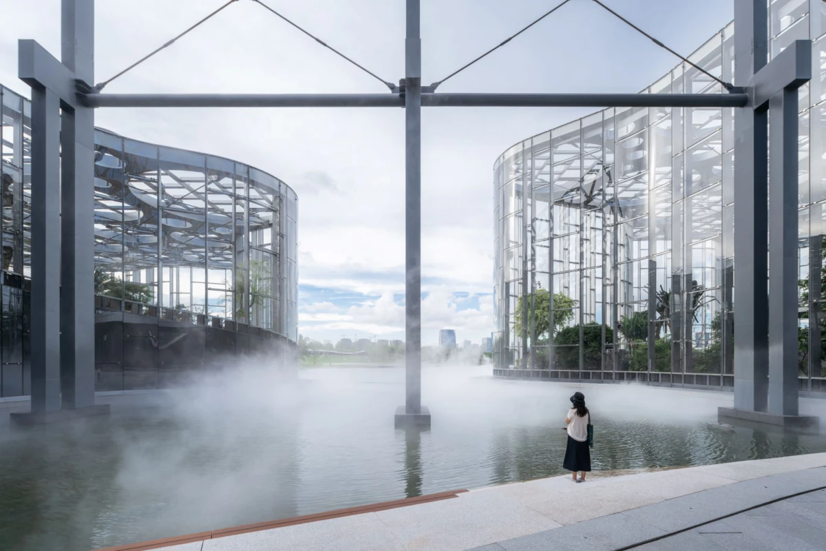Person standing at the edge of a misty reflecting pool between glass-walled modern buildings