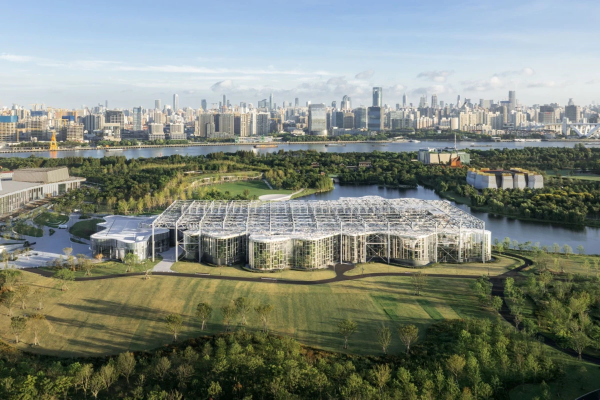 Curved glass building in a landscaped park by a lake, with a distant city skyline.