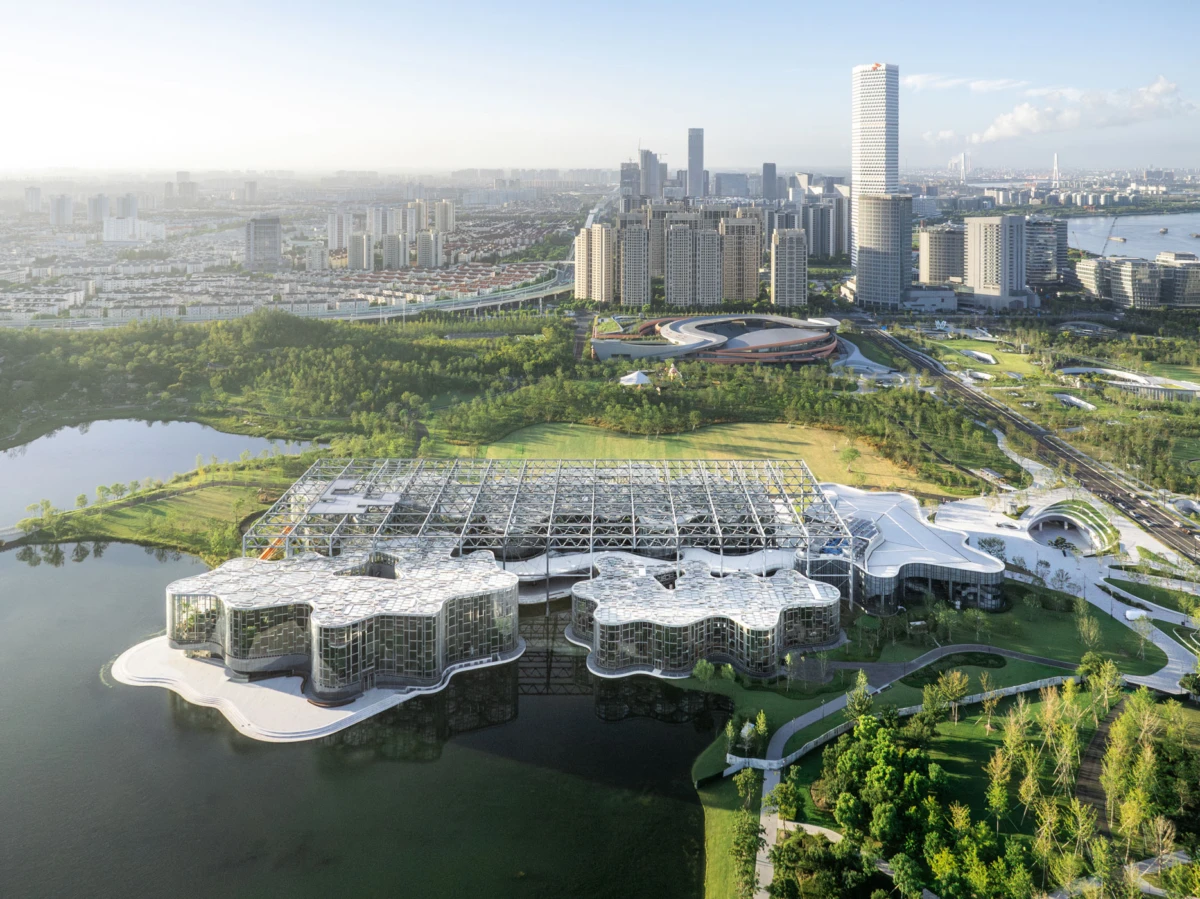 Aerial view of a futuristic glass-walled building complex on a lake, connected by curved white walkways, with parks and a distant skyline.
