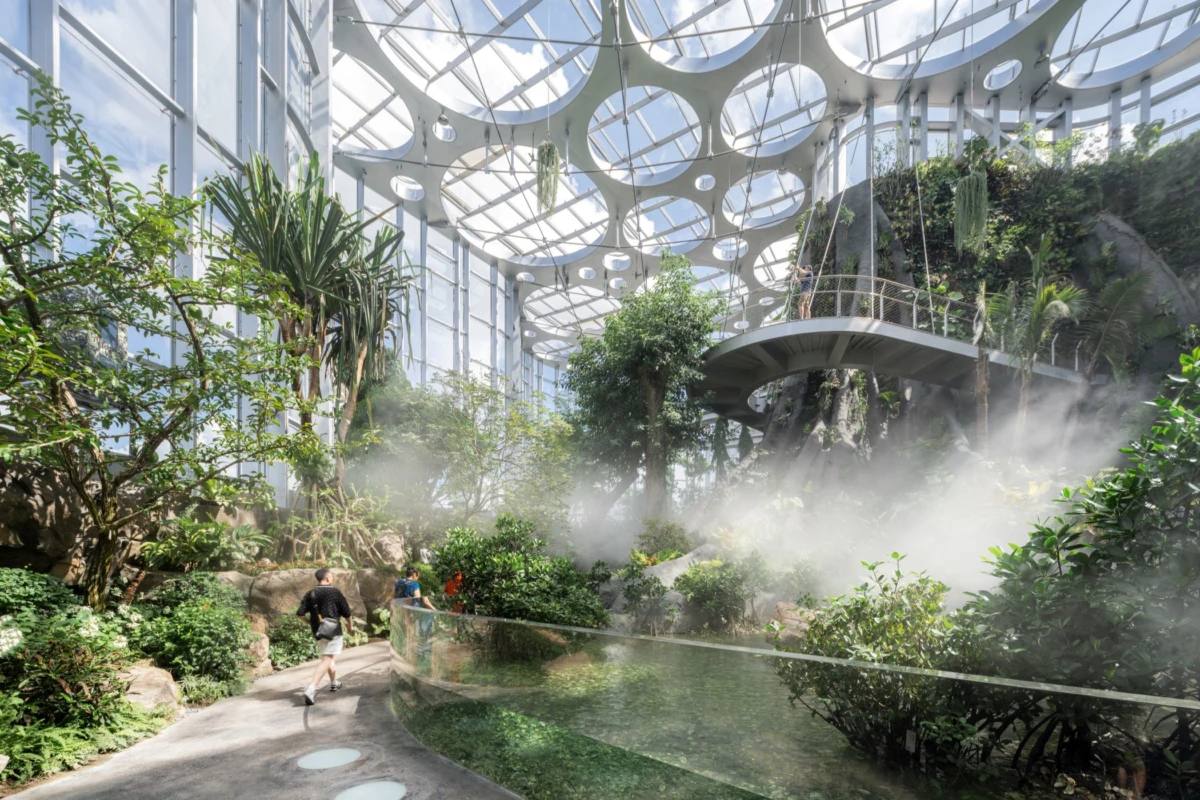 Indoor glass-domed conservatory with tropical plants, misty pond, and curved elevated walkway; two people stroll along a path.