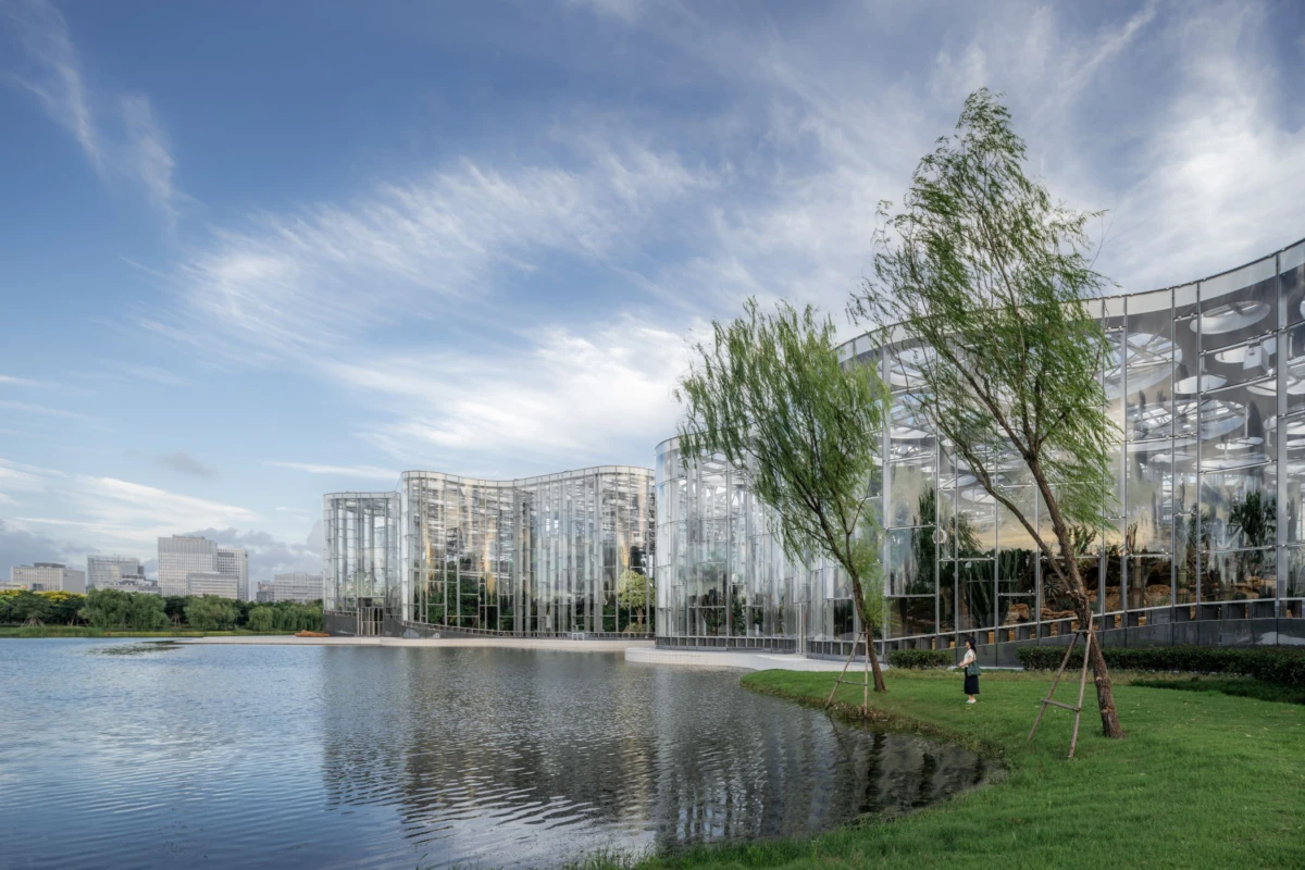 Glass-clad modern building along a lakefront; a person walks on the grassy bank beside slender trees.