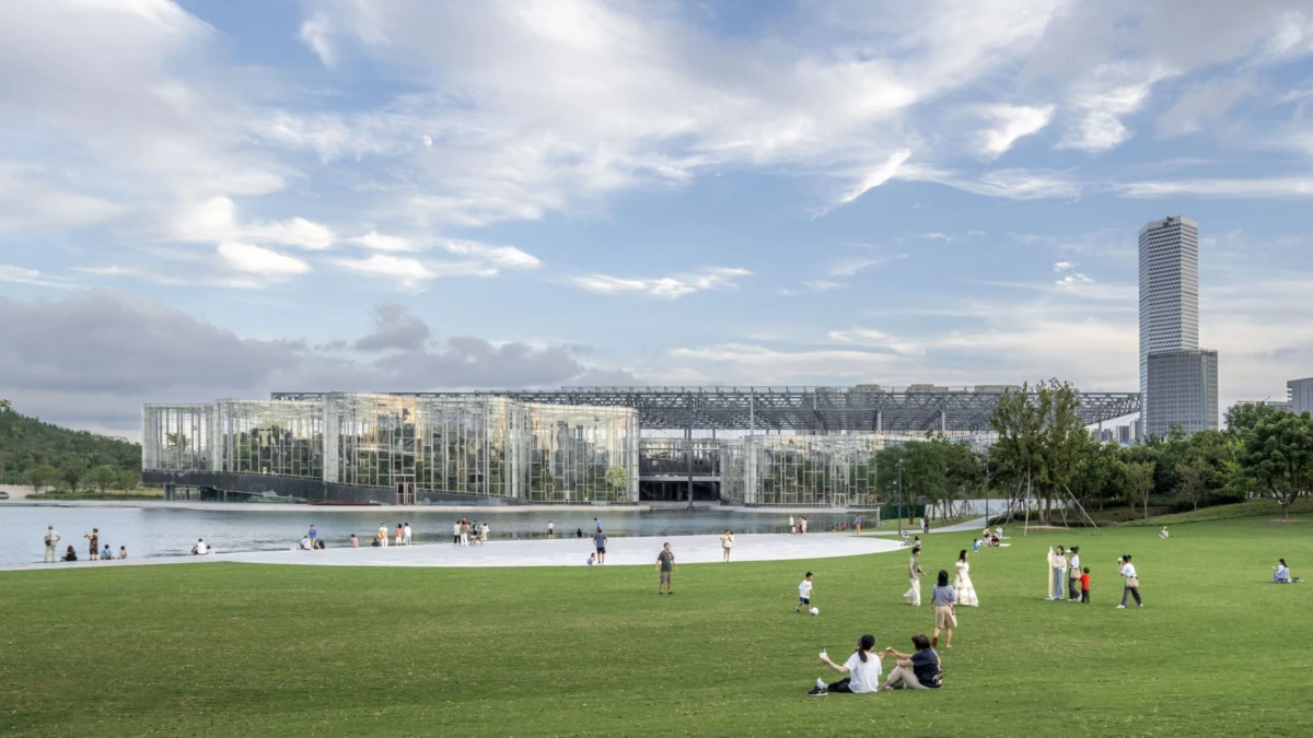 Public park by a lake with a glass-walled modern building; people walk and sit on the grass as a tall skyscraper looms in the distance.