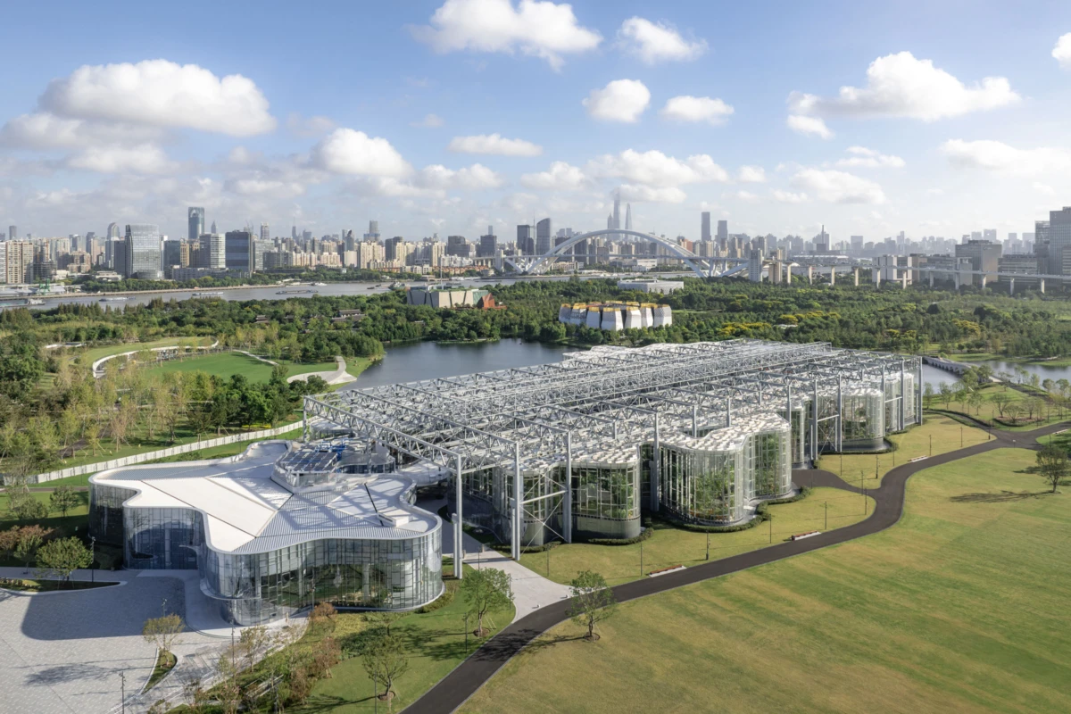 Futuristic glass-and-metal building complex with connected cylindrical modules in a park along a river; urban skyline and arch bridge in background.