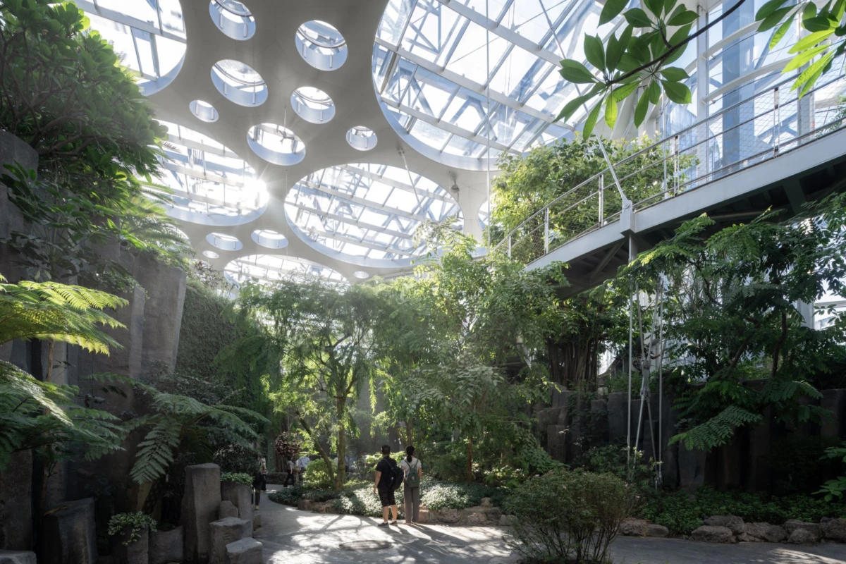 Two visitors walk through a sunlit indoor botanical conservatory with glass skylights and lush greenery.