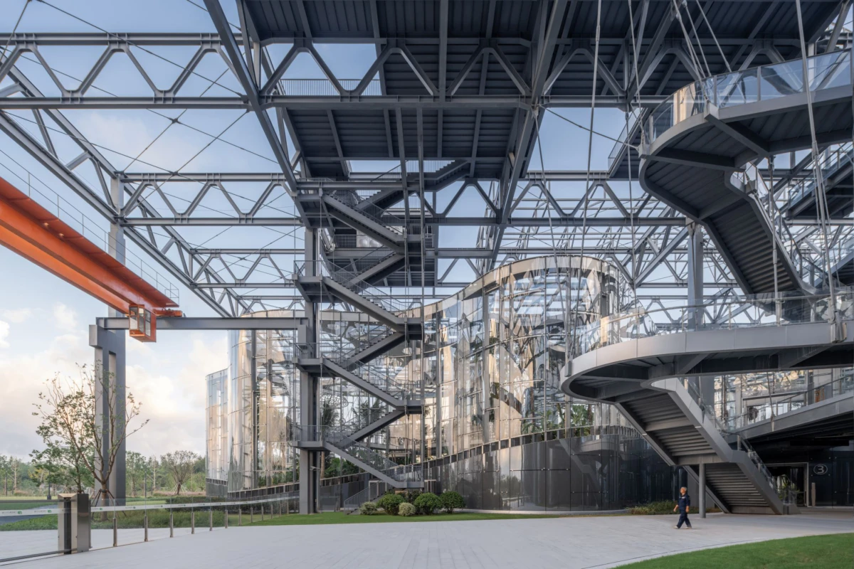 Modern glass-and-steel building with exposed metal framework and exterior spiral staircases; a person walks at the outdoor base.