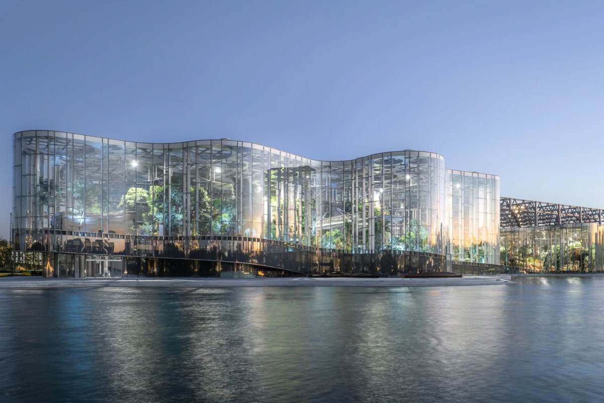 Glass-walled conservatory on the waterfront at dusk, interior greenery visible through curved glass.