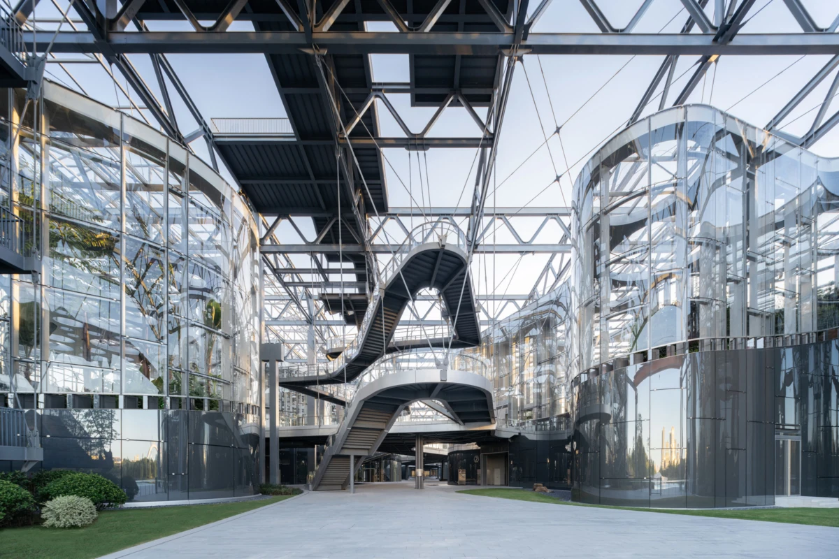 Modern glass atrium with curved, suspended steel stairs and elevated walkways between glass-walled buildings.