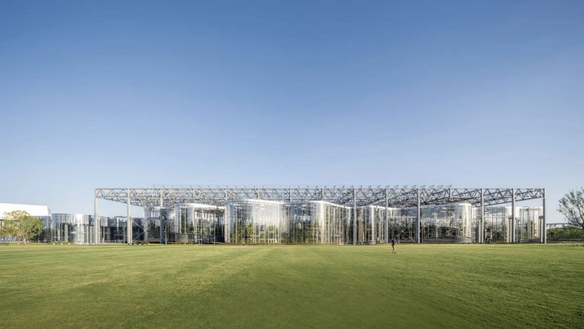 Modern glass-and-steel building with lattice frame behind a wide green lawn; a person walks across the field under a clear blue sky.