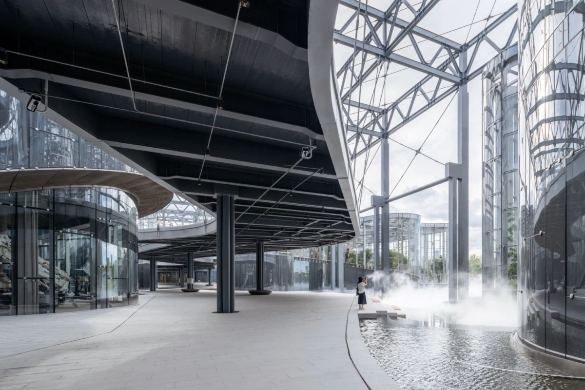 Modern glass-and-steel complex with an elevated curved walkway; a person stands beside a misty water feature.