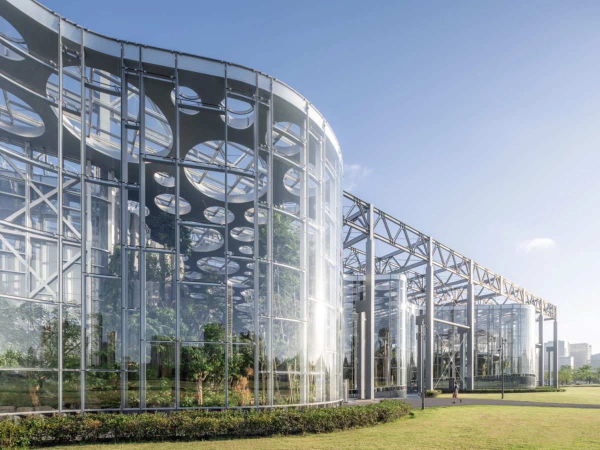 Modern glass conservatory with curved cylindrical facade and steel framework; a person walking on the manicured lawn nearby.