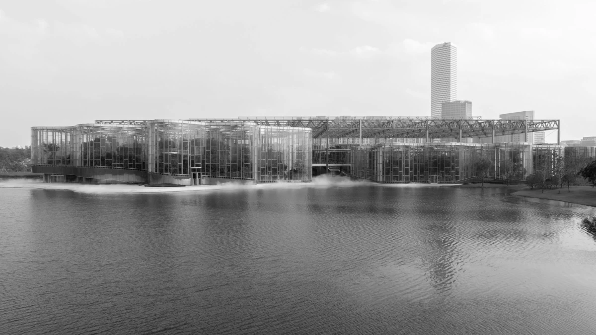 Glass-fronted, industrial-style building along a calm river; reflections on water with a tall rectangular skyscraper in the background.