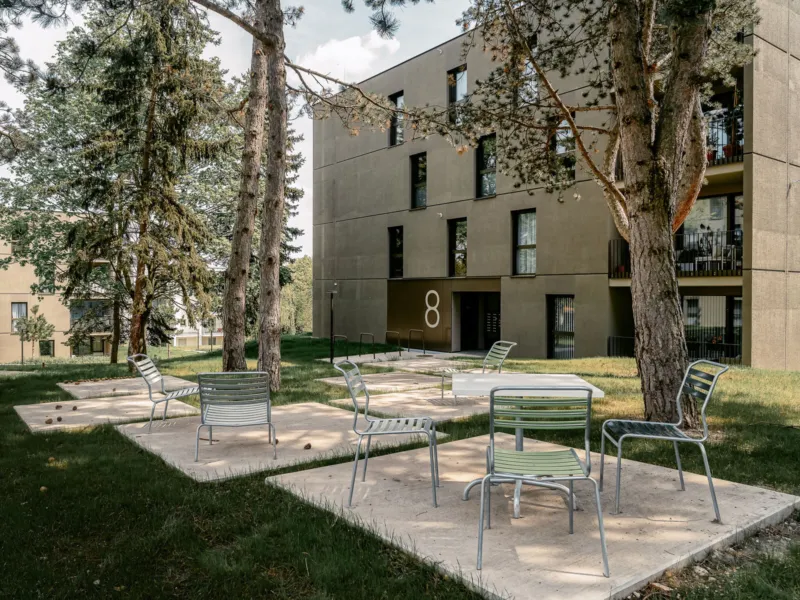 Outdoor seating area in a residential courtyard with metal chairs on concrete platforms, beige apartment building and trees in the background.