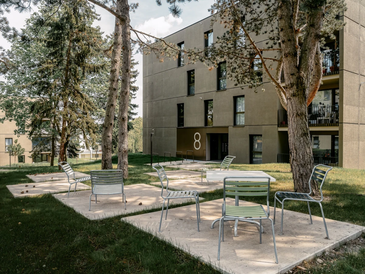 Outdoor seating area in a residential courtyard with metal chairs on concrete platforms, beige apartment building and trees in the background.