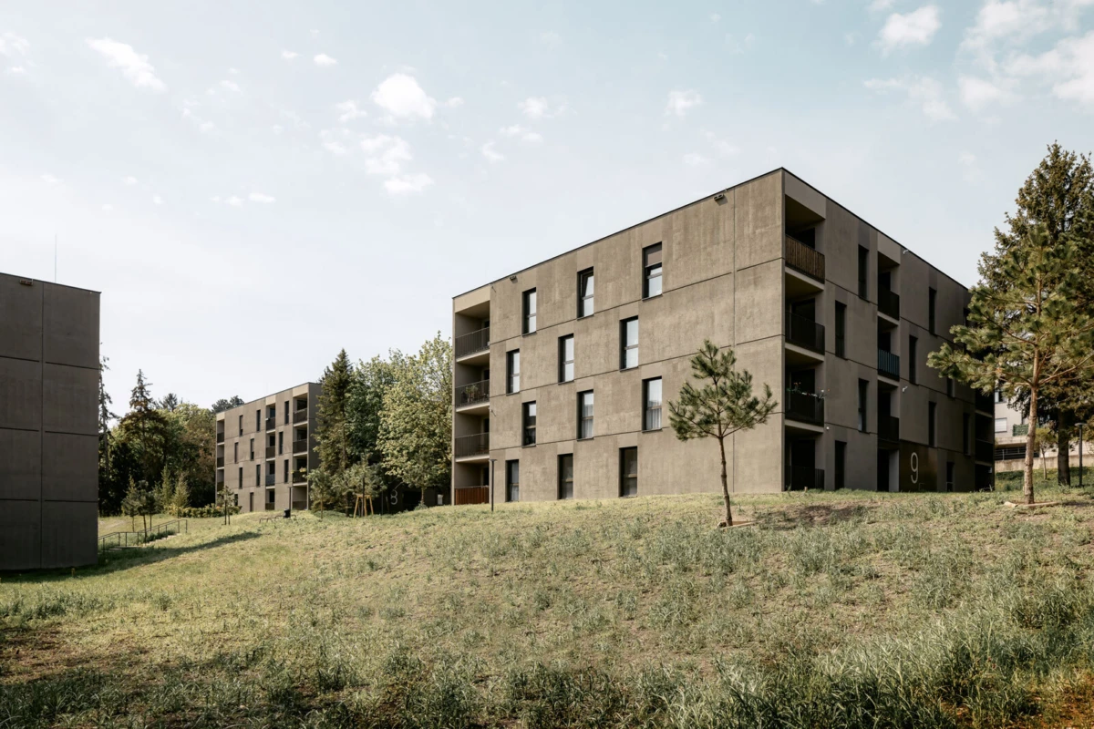 Exterior view of a modern concrete apartment complex on a grassy field with scattered trees under a blue sky.