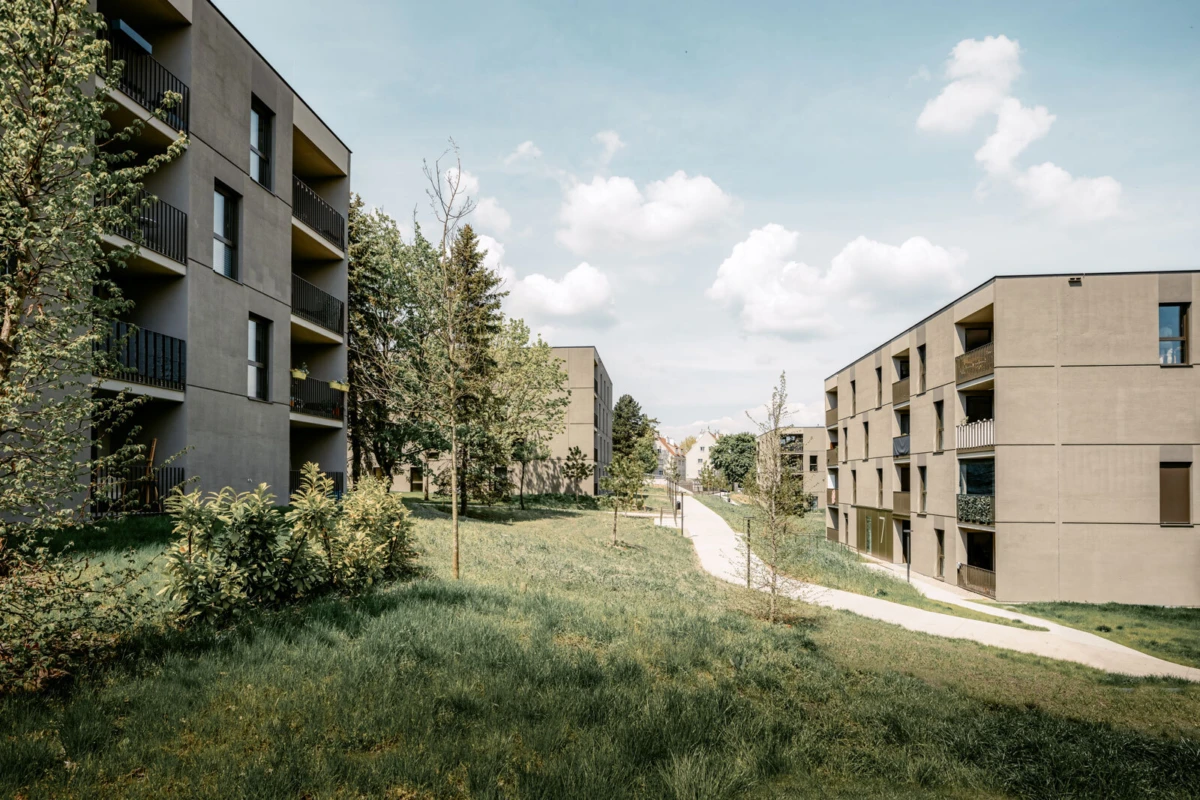 Beige apartment blocks flank a grassy courtyard with a winding path, trees, and shrubs under a blue sky.