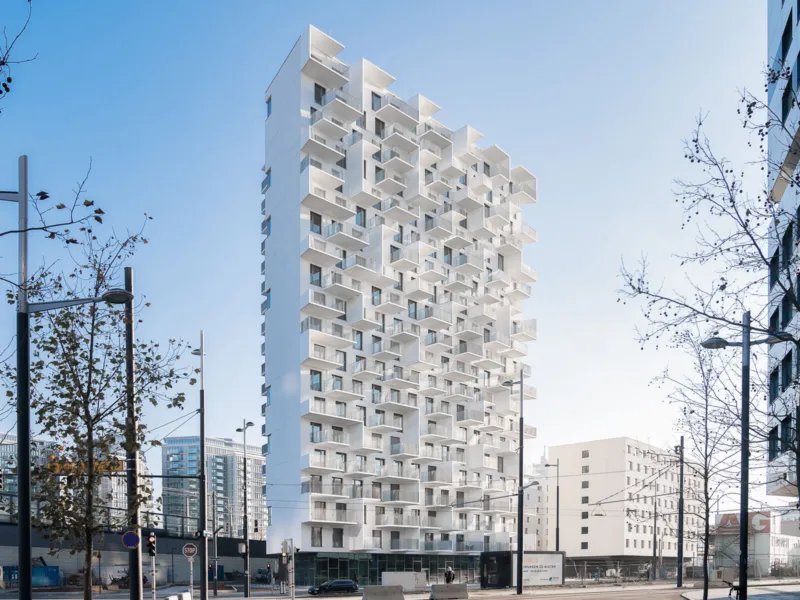 Tall white apartment tower with stacked balconies on an urban street; leafless trees and surrounding buildings.