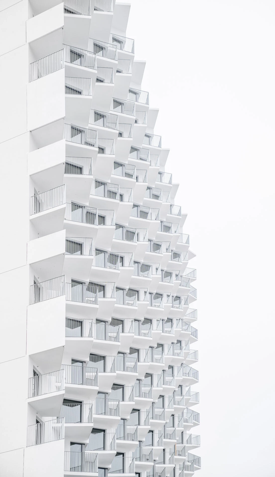 Tall white apartment building with repeating protruding balconies forming a diagonal facade against a pale sky.