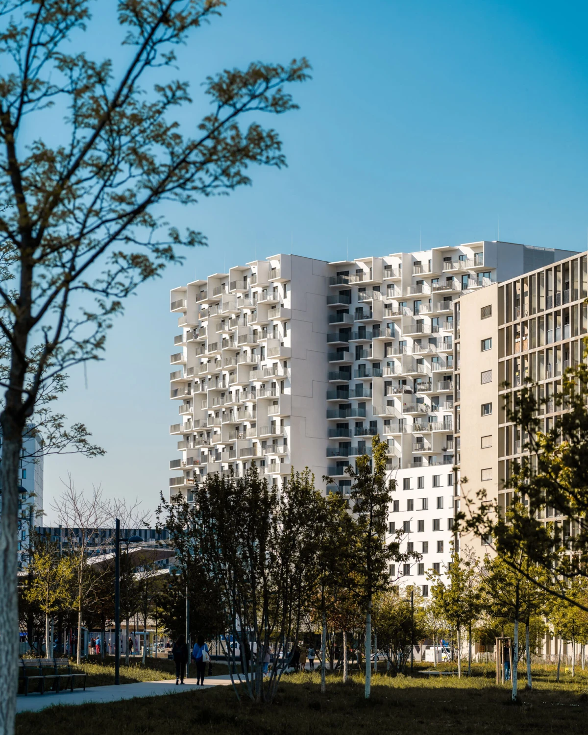 Apartment building with stacked cantilevered balconies in a tree-lined park; pedestrians walk along a path.