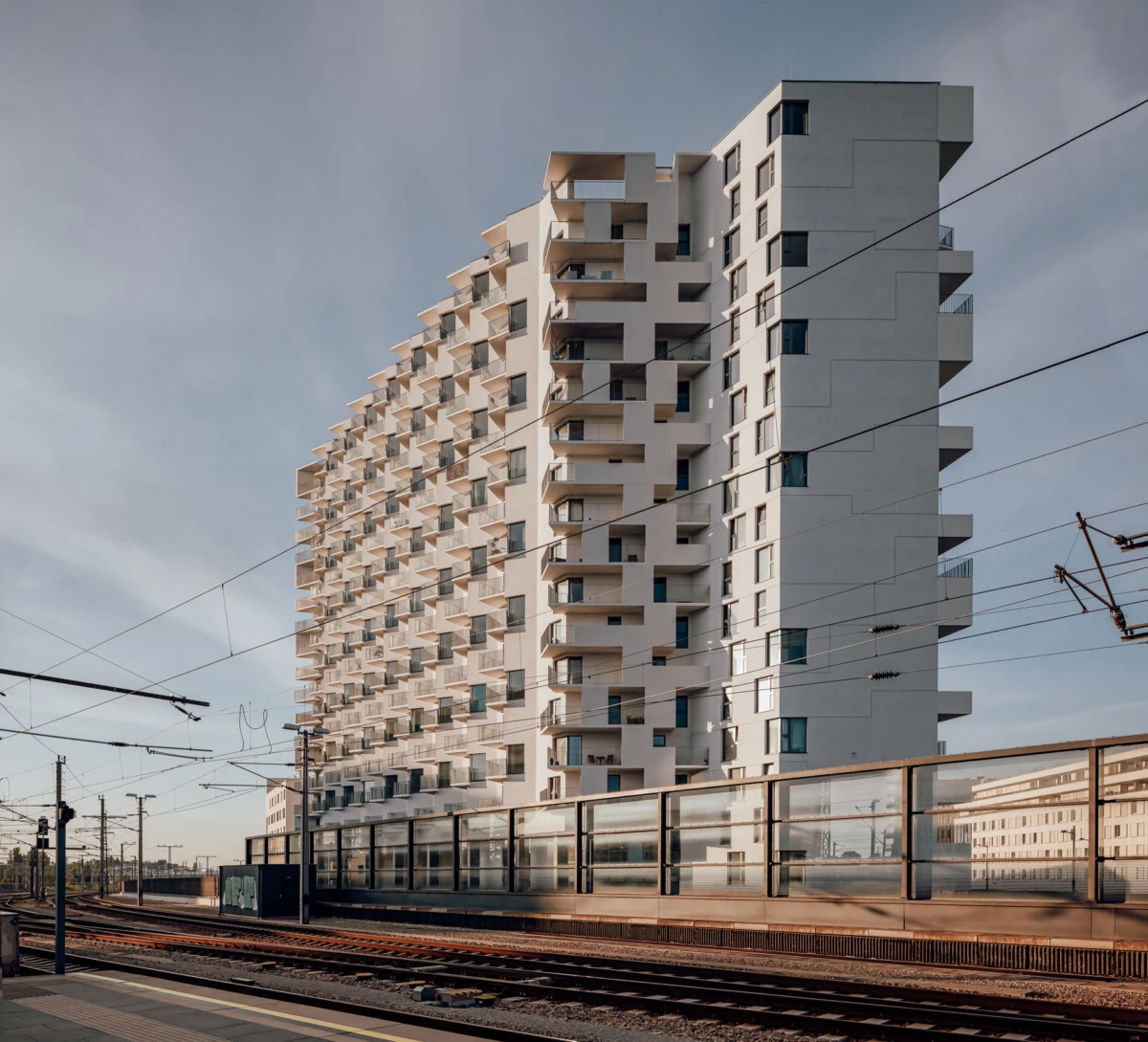 High-rise apartment building with staggered balconies beside railway tracks and a glass-walled platform barrier.