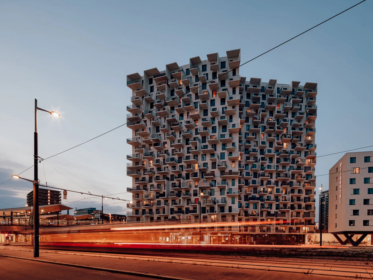 Apartment building with stacked balconies forming a honeycomb facade, red tram light trails along a city street at dusk.