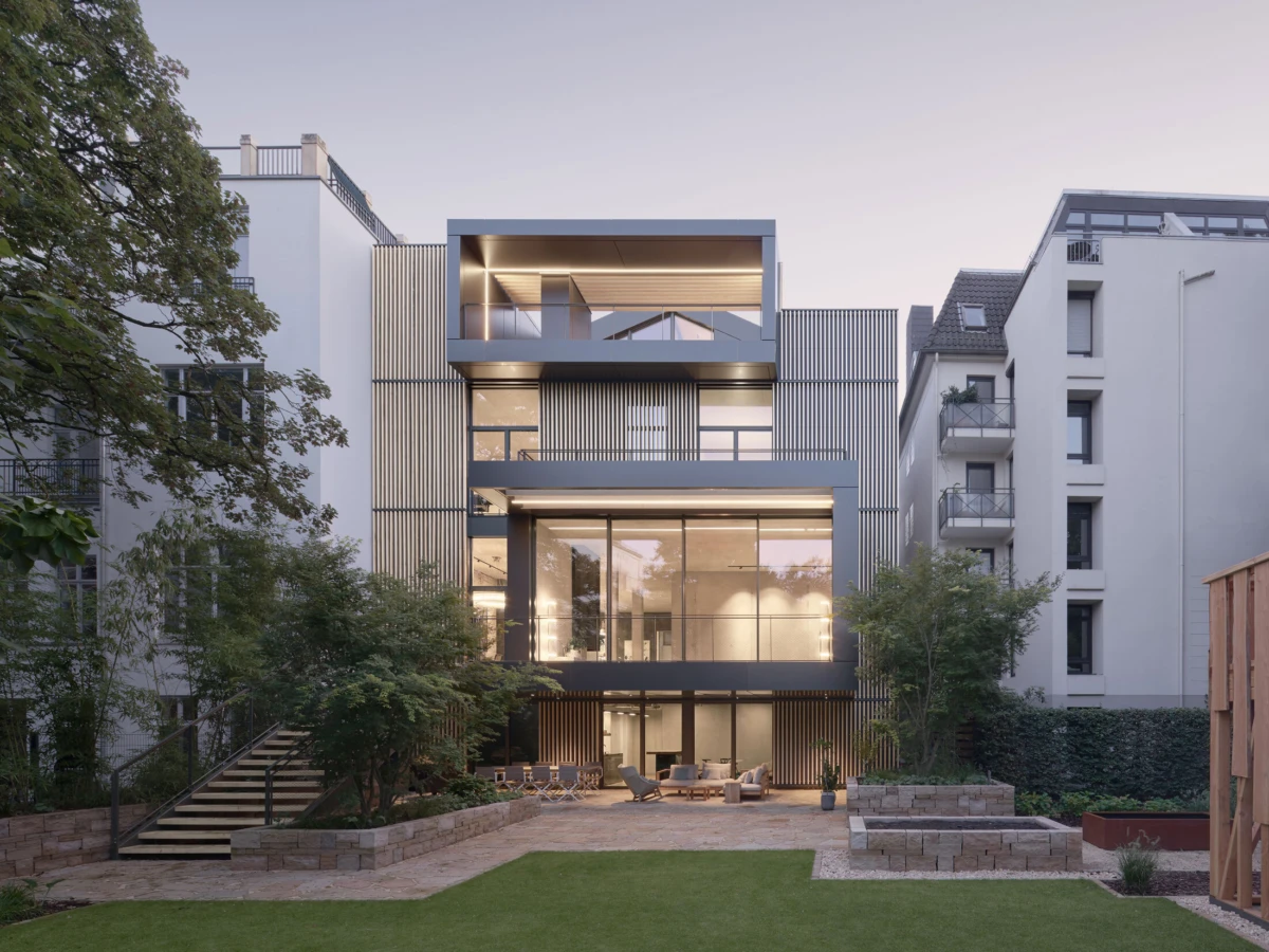 Modern glass-fronted apartment building with terraces, illuminated interiors, in a landscaped courtyard with trees and adjacent white buildings.