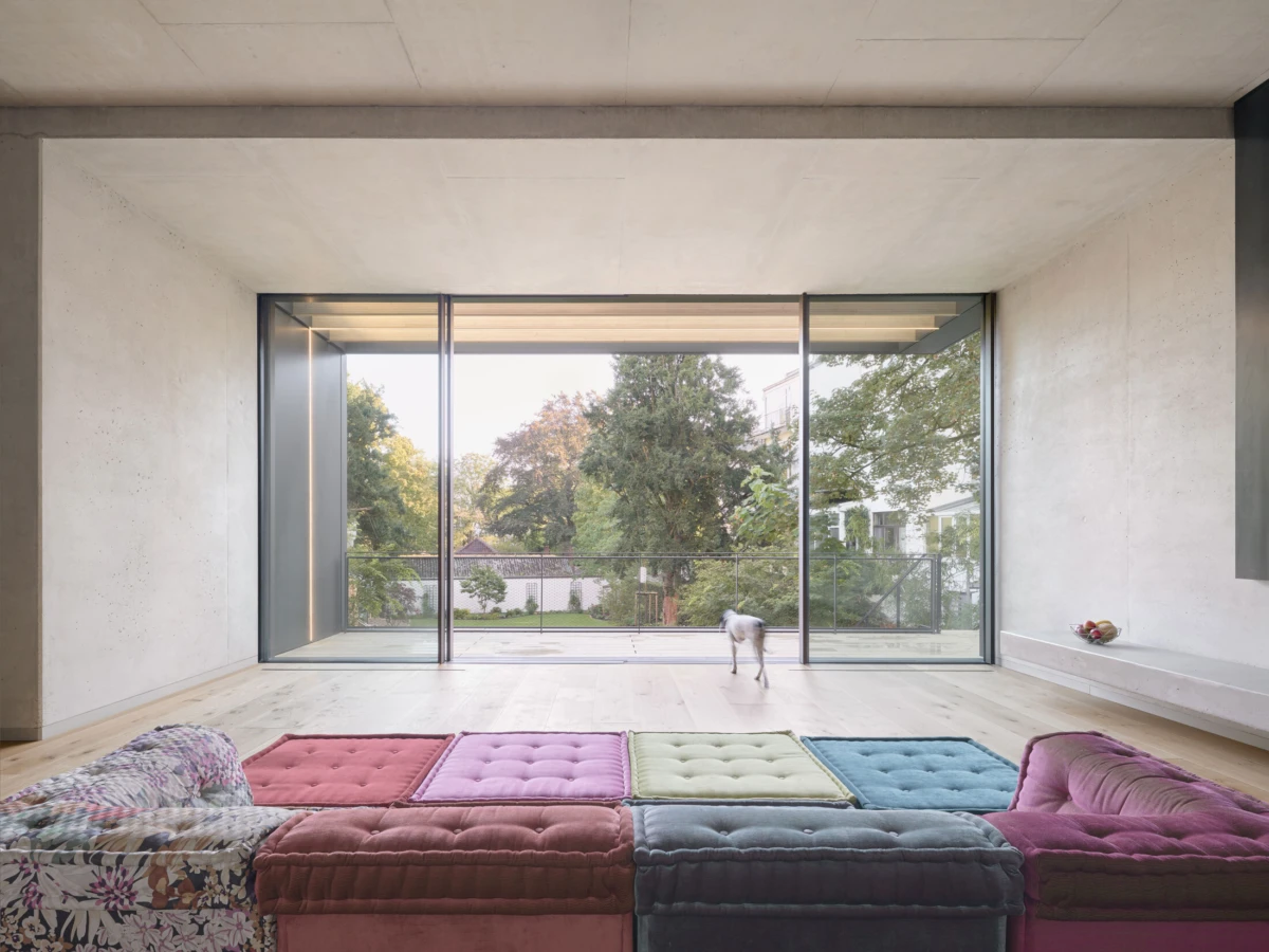 Small dog walking toward the glass wall in a modern living room with colorful tufted ottomans and a garden outside.