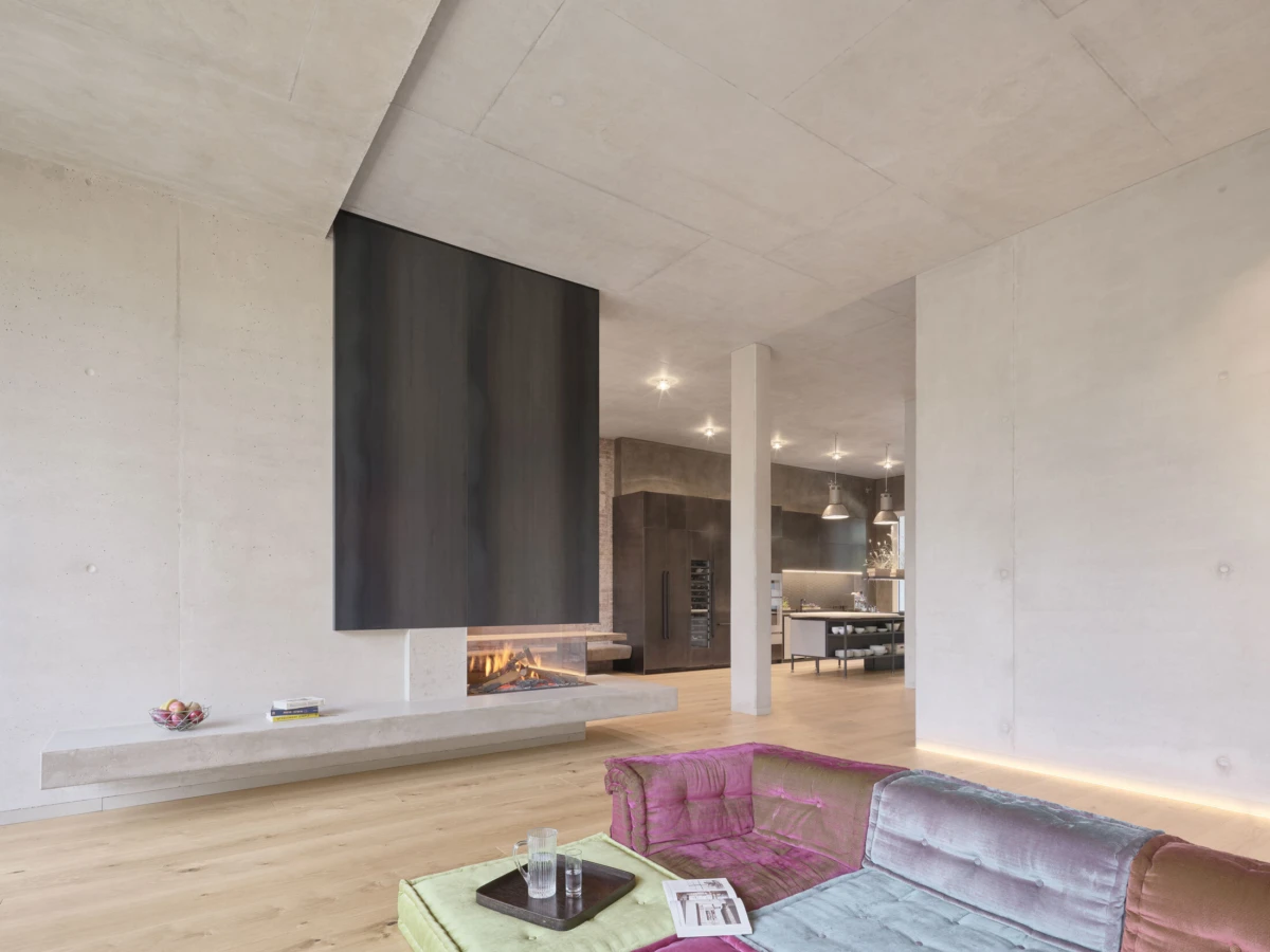 Contemporary open-plan living room: concrete walls and ceiling, fireplace with dark panel, pastel velvet sofa, and open kitchen in the background.