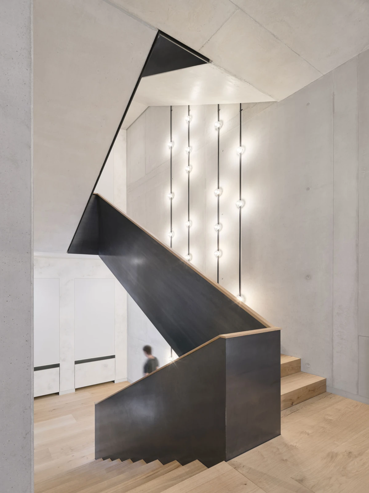 Modern concrete stairwell with a black metal staircase and wooden treads; row of hanging globe lights on the wall, a blurred figure descends.