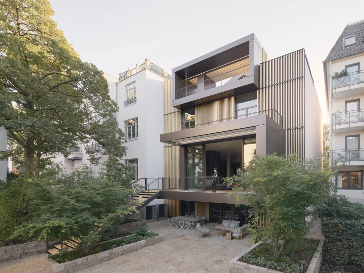 Contemporary multi-story apartment building with dark brown panels; person on the second-floor balcony; stone courtyard with seating and trees.