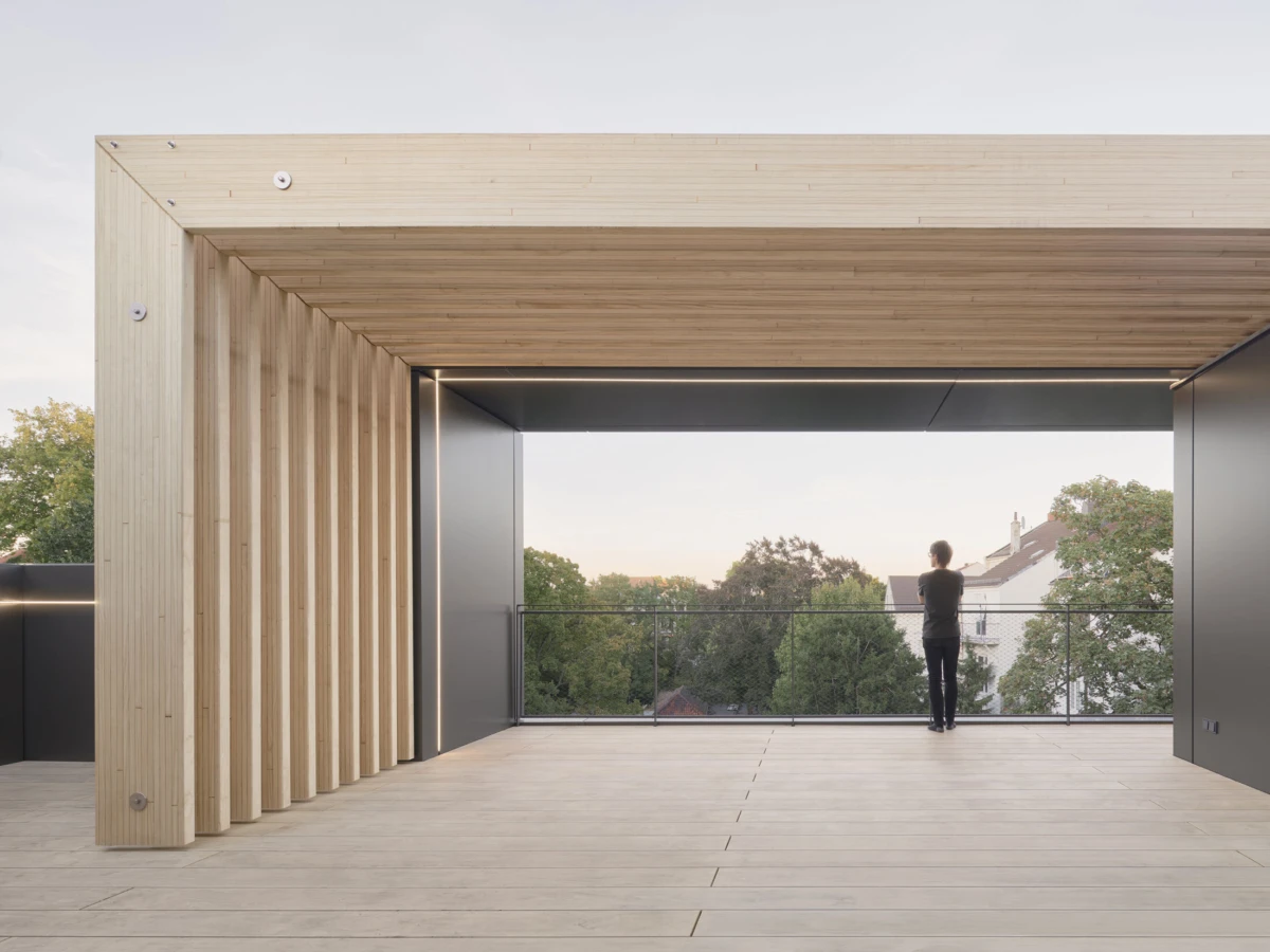 Person stands at the glass railing of a modern wooden terrace, looking out over trees and rooftops.