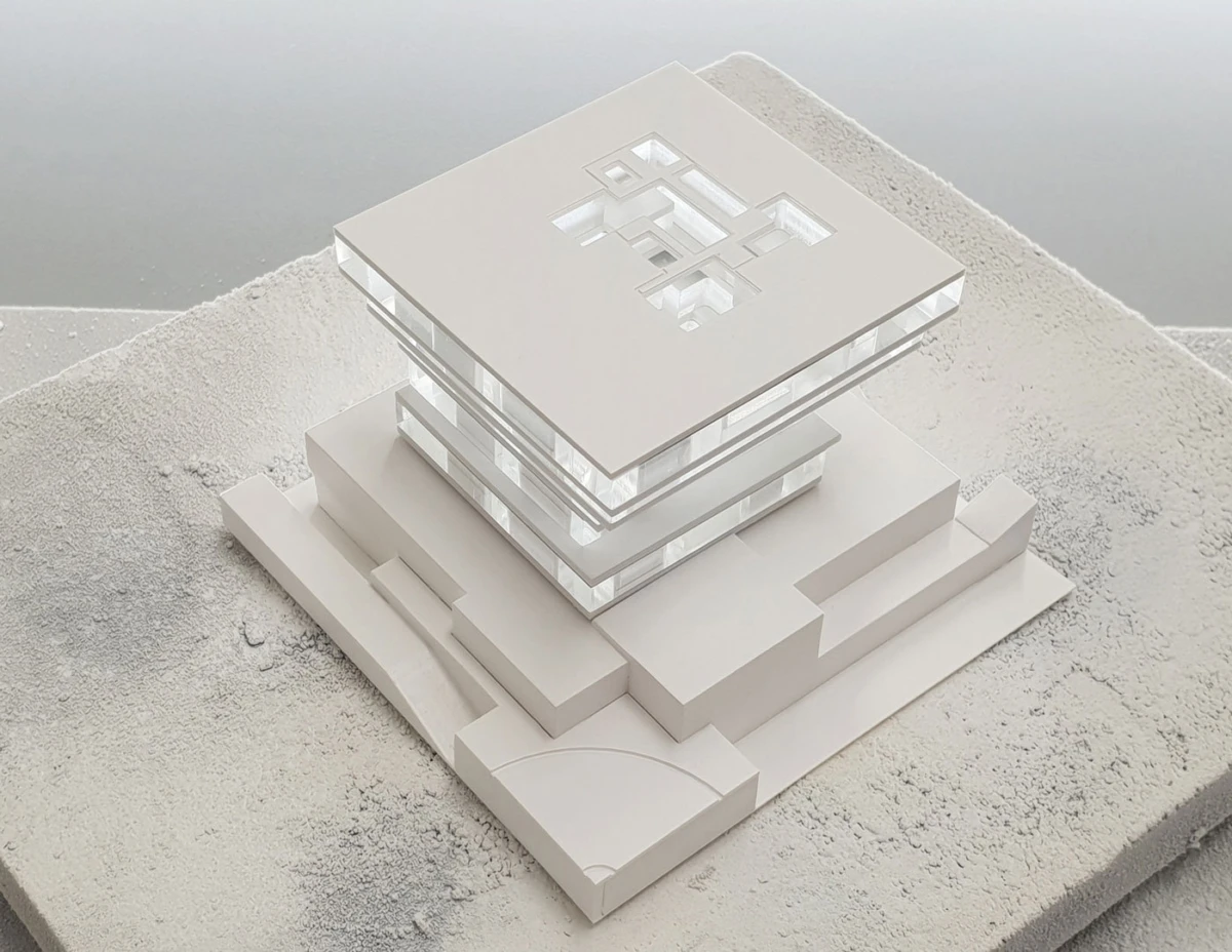 White architectural model of a multi-level glass building with a square open roof, placed on a textured sandy base.