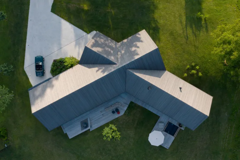 An aerial view of a wooden-roofed, multi-wing house on a green lawn; a car is parked in the driveway.